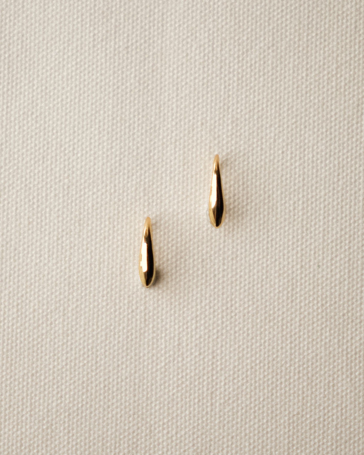 Two small gold orzo pasta-like earrings on a textured cream-white fabric background.