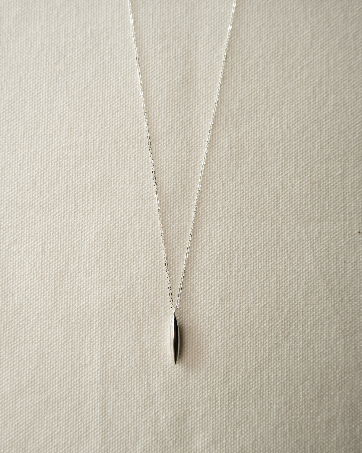 A delicate silver chain necklace with a small, polished silver grain-like pendant, displayed on a textured beige fabric background.