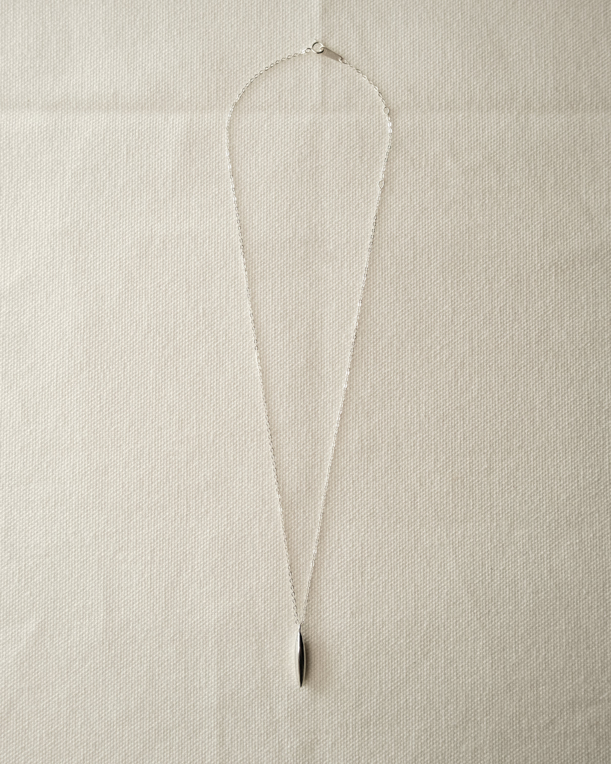 A delicate silver chain necklace with a small, polished gold grain-like pendant, laid flat on a textured off-white fabric background.