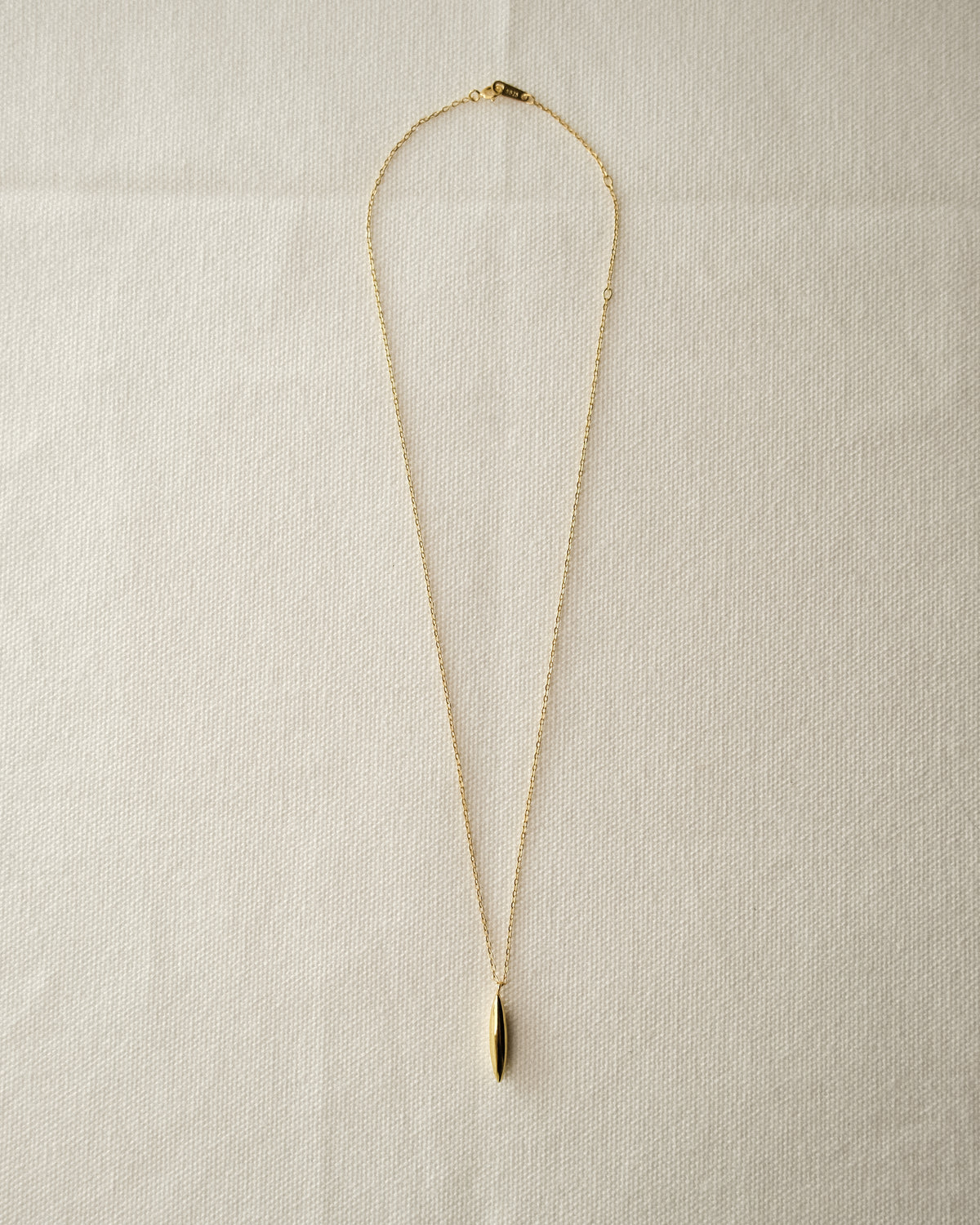 A delicate gold chain necklace with a small, polished gold grain-like pendant, laid flat on a textured off-white fabric background.