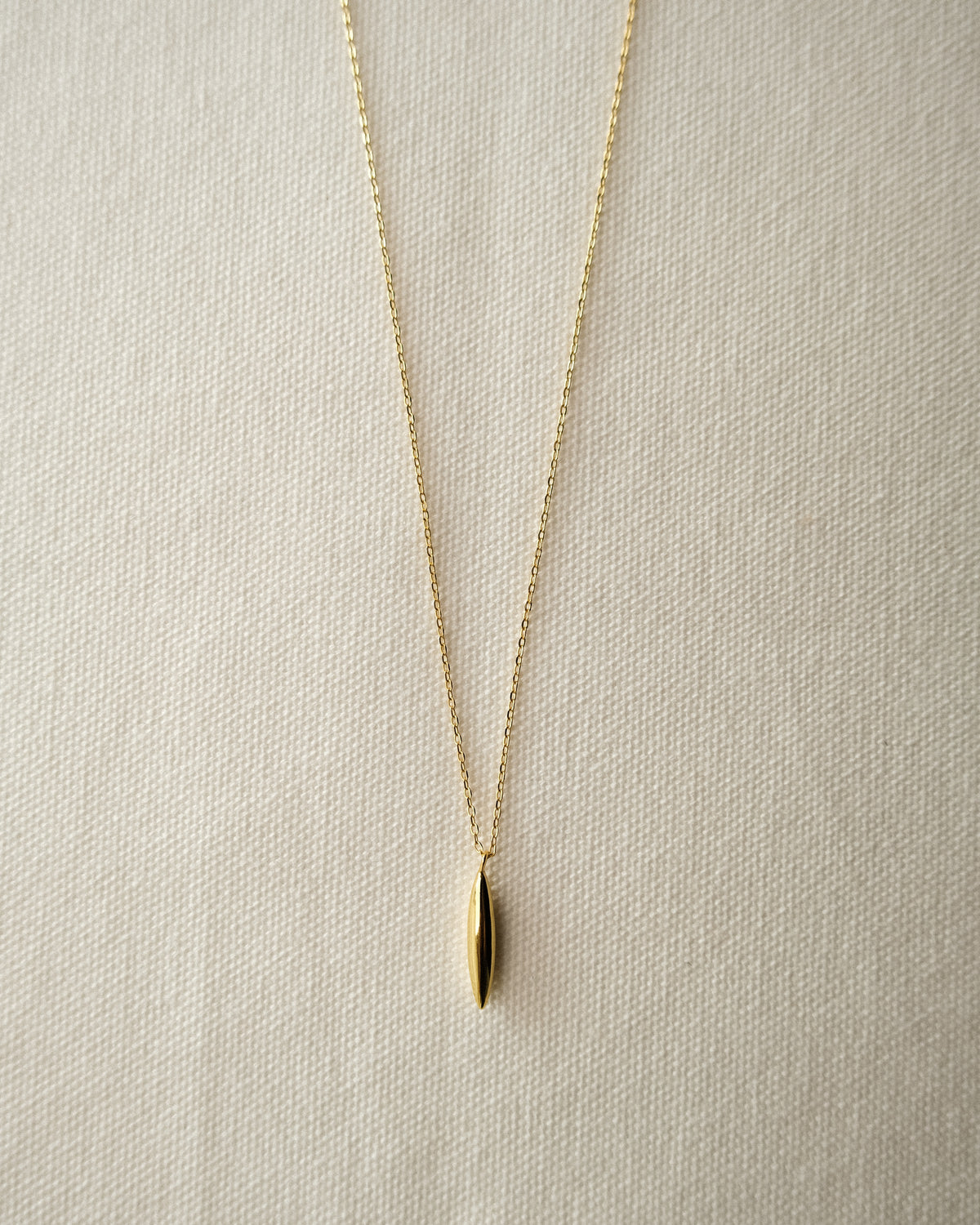 A delicate gold chain necklace with a small, vertically grain-like pendant, displayed on a textured cream fabric background.