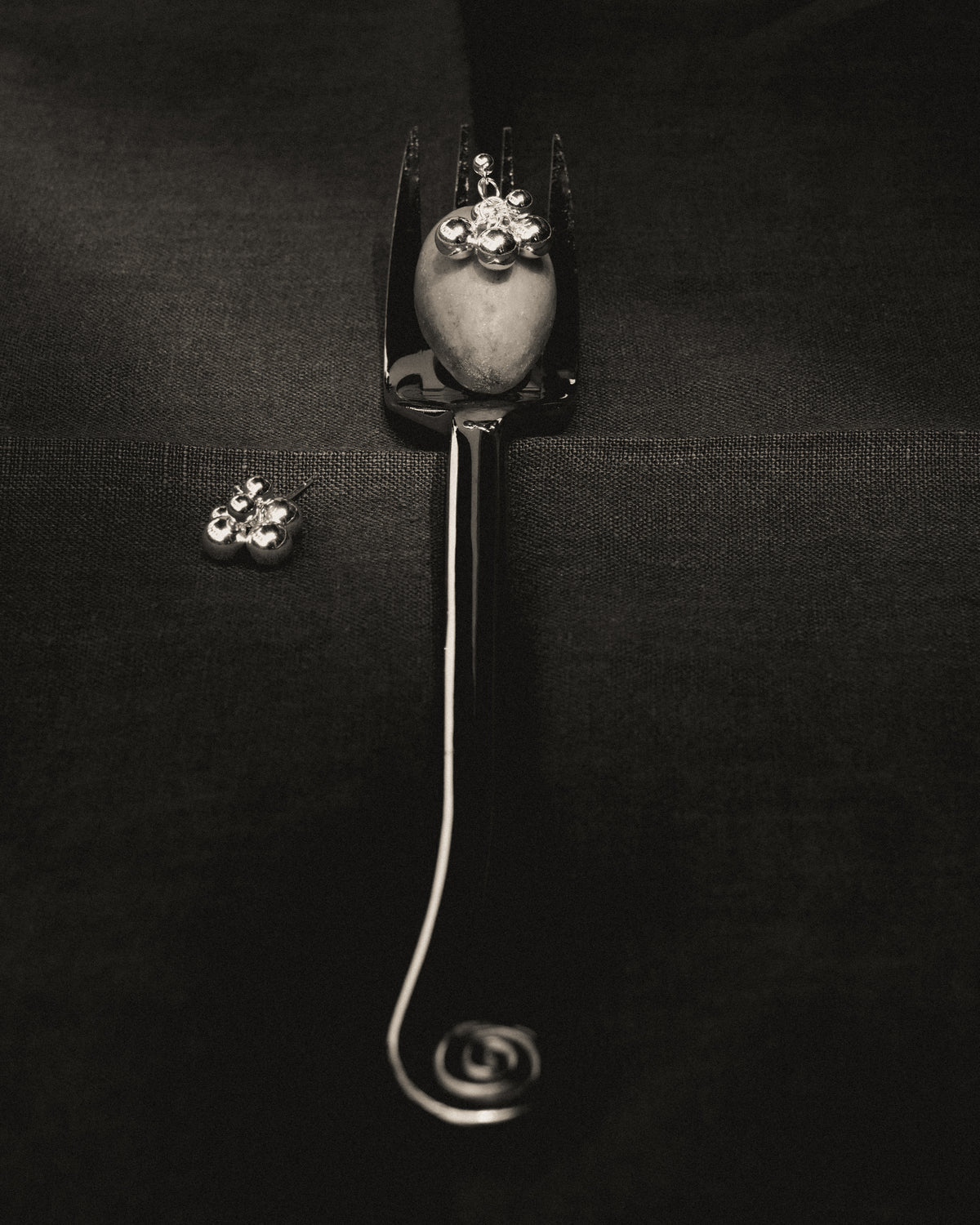 Black and white photograph of silver grape cluster stud earrings, one perched on an olive atop a fork with a spiral handle, the other resting nearby on dark textured fabric.