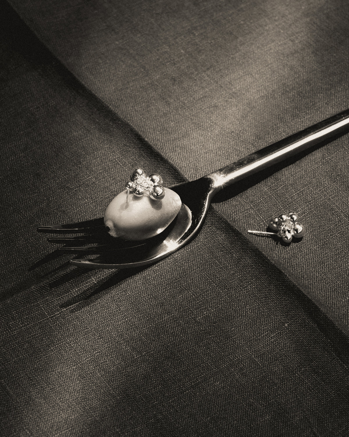 Black and white close up of silver grape cluster stud earrings, one resting on an olive atop a metal fork and the other on dark fabric, illuminated by natural light.