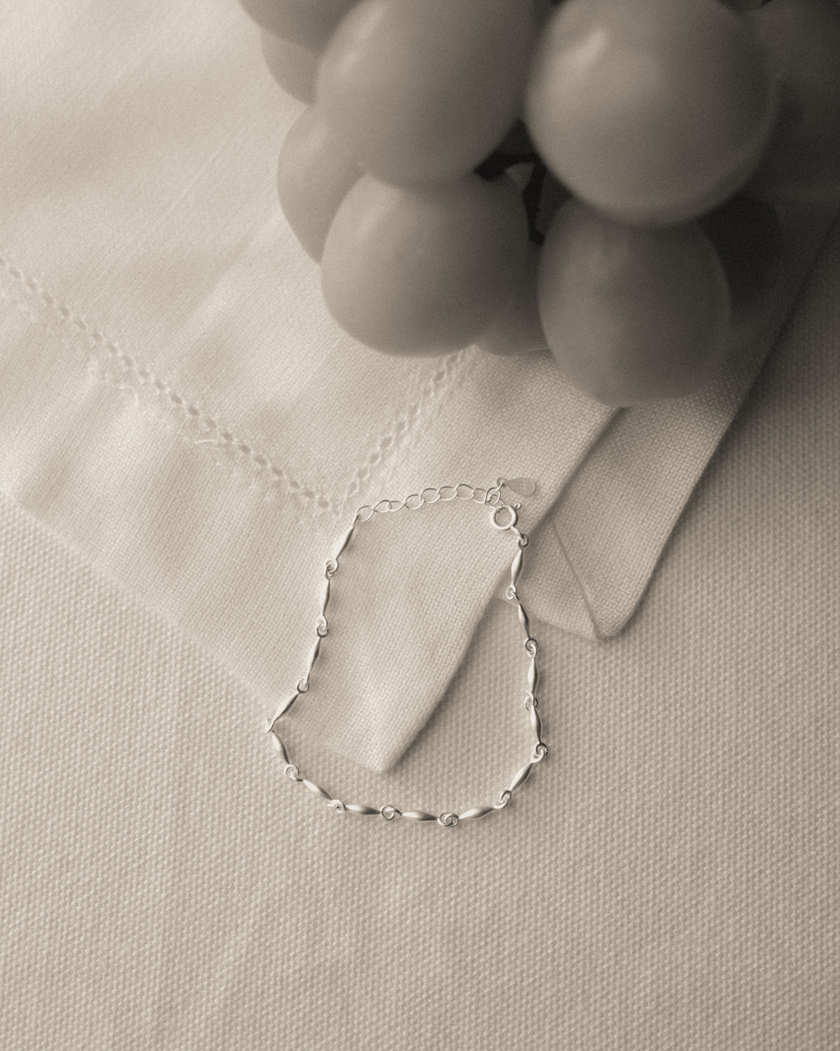 A delicate silver chain bracelet with small bead-like links, laid on a textured white fabric surface next to a white napkin and a blurred gray decorative object.