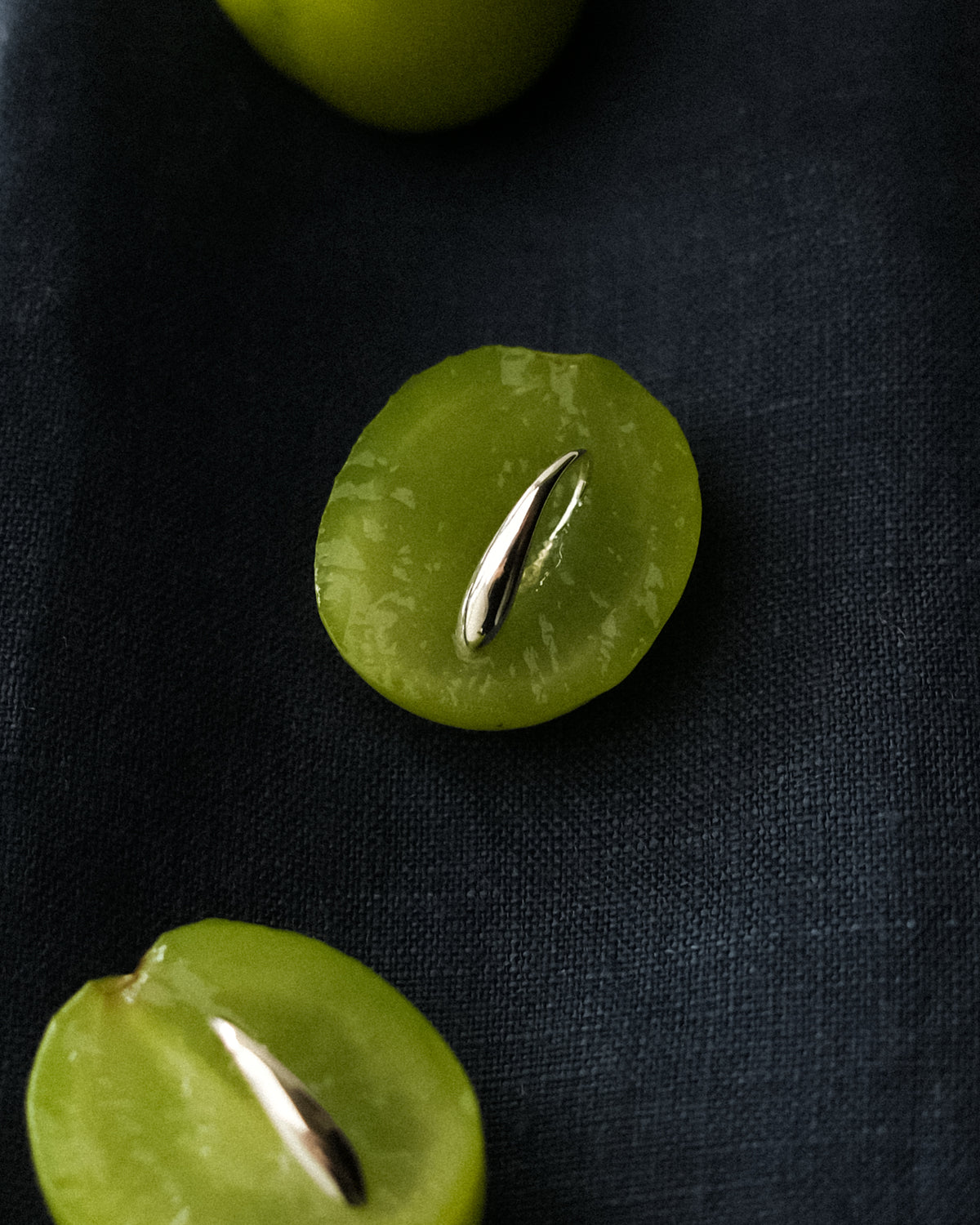 A close-up of a silver slip-on earring, placed on a sliced green grape against a dark navy textured fabric background.