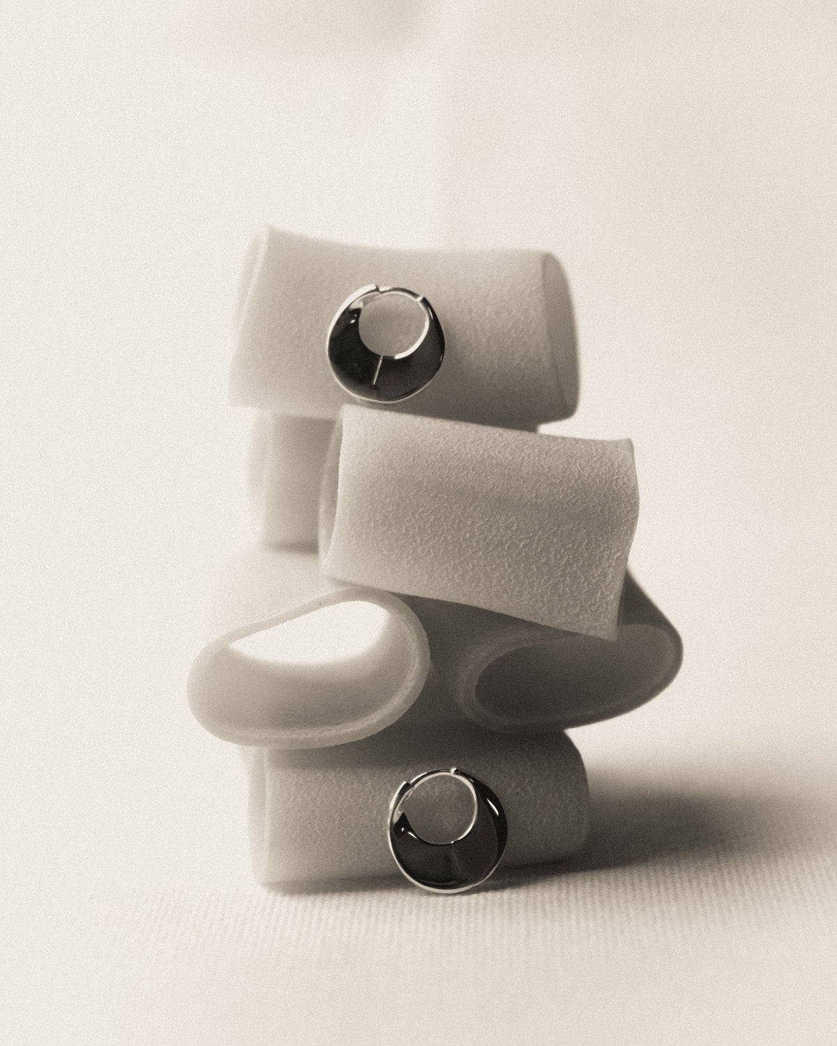 A few paccheri pasta arranged in a stacked, abstract composition with a pair of scupltural hoop earrings standing or resting on top, on a warm off-white background. 