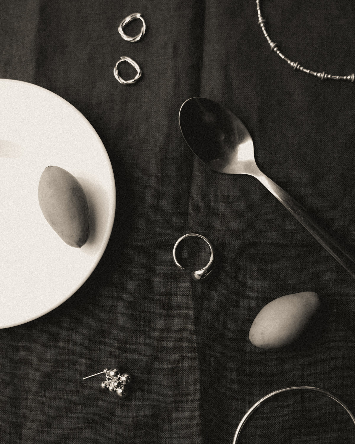 A styled flat-lay of silver-toned jewelry and silverware on a dark textured fabric background, including rings, a necklace, a spoon, a bracelet, and a twisted hoop earring, with two olive resting on a white plate and another on the dark textured fabric.