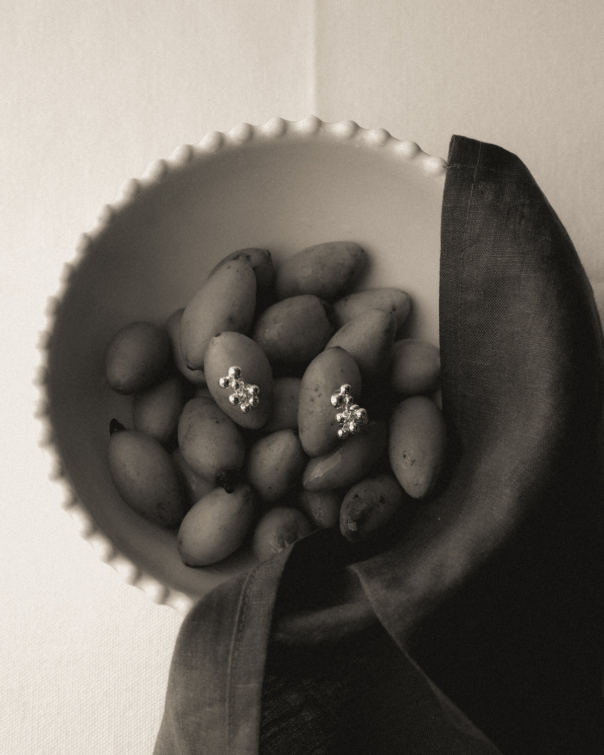 A pair of silver grape cluster stud earrings resting on olives in a white scalloped bowl, with dark fabric draped over the rim against a neutral background.