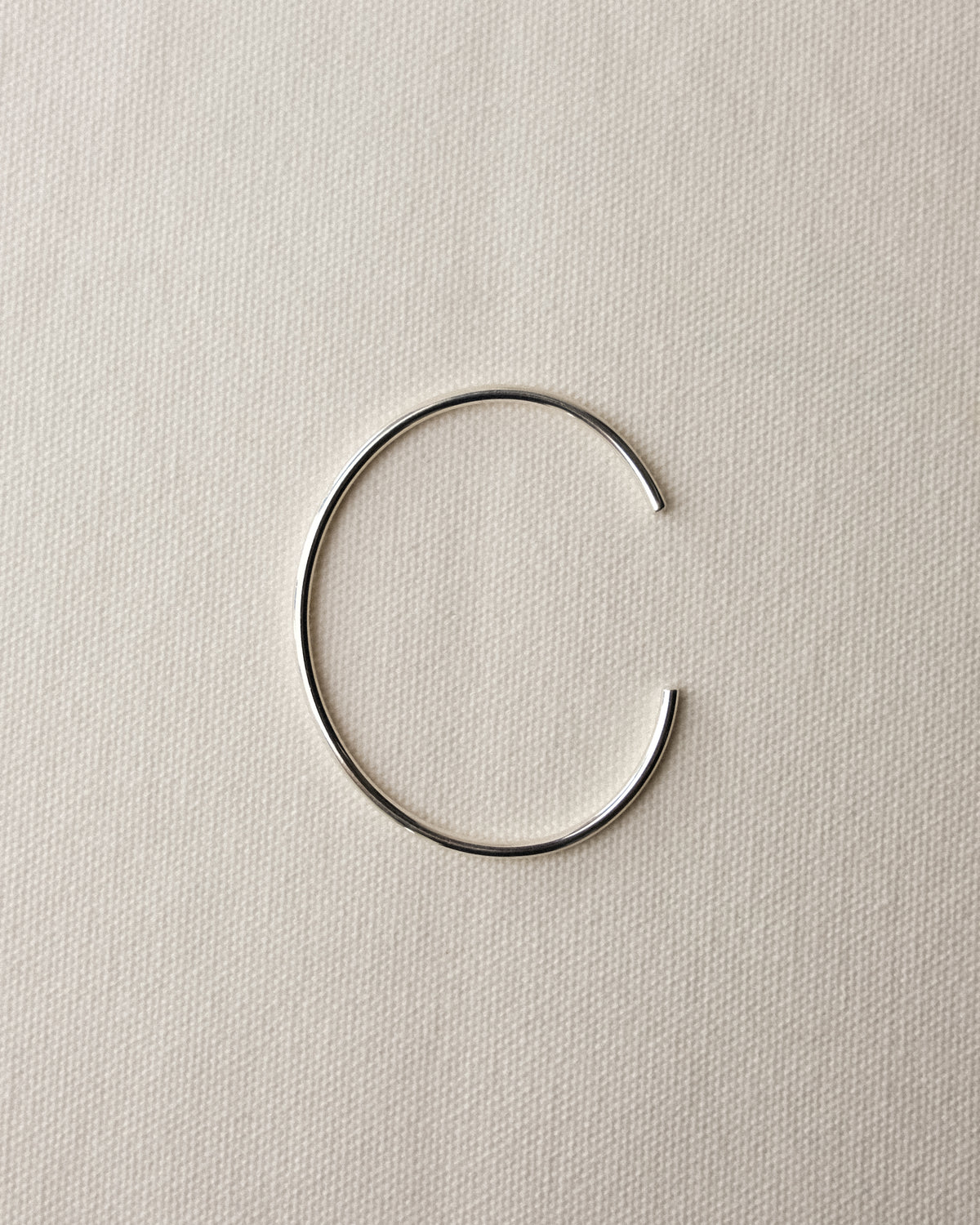 A minimalist, open circular cuff bracelet made of a thin sterling silver, displayed on a textured cream-white fabric background.