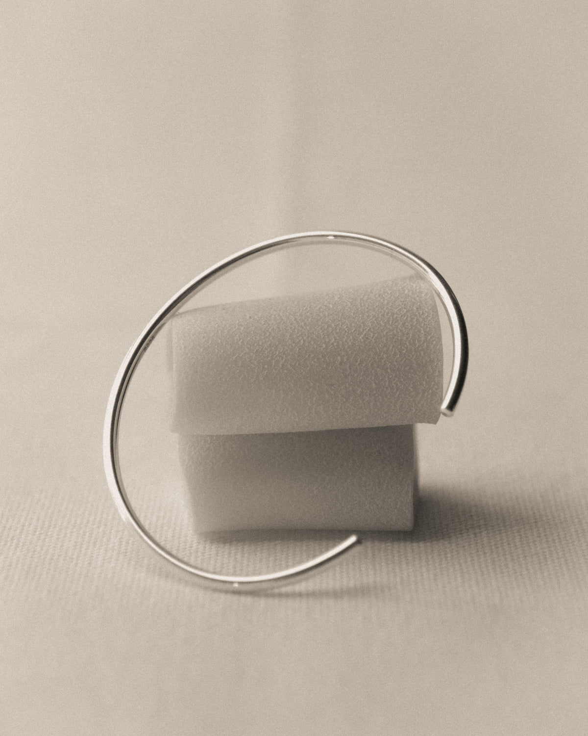 A minimalist silver open cuff bracelet displayed on 2 paccheri pasta against a neutral beige background.