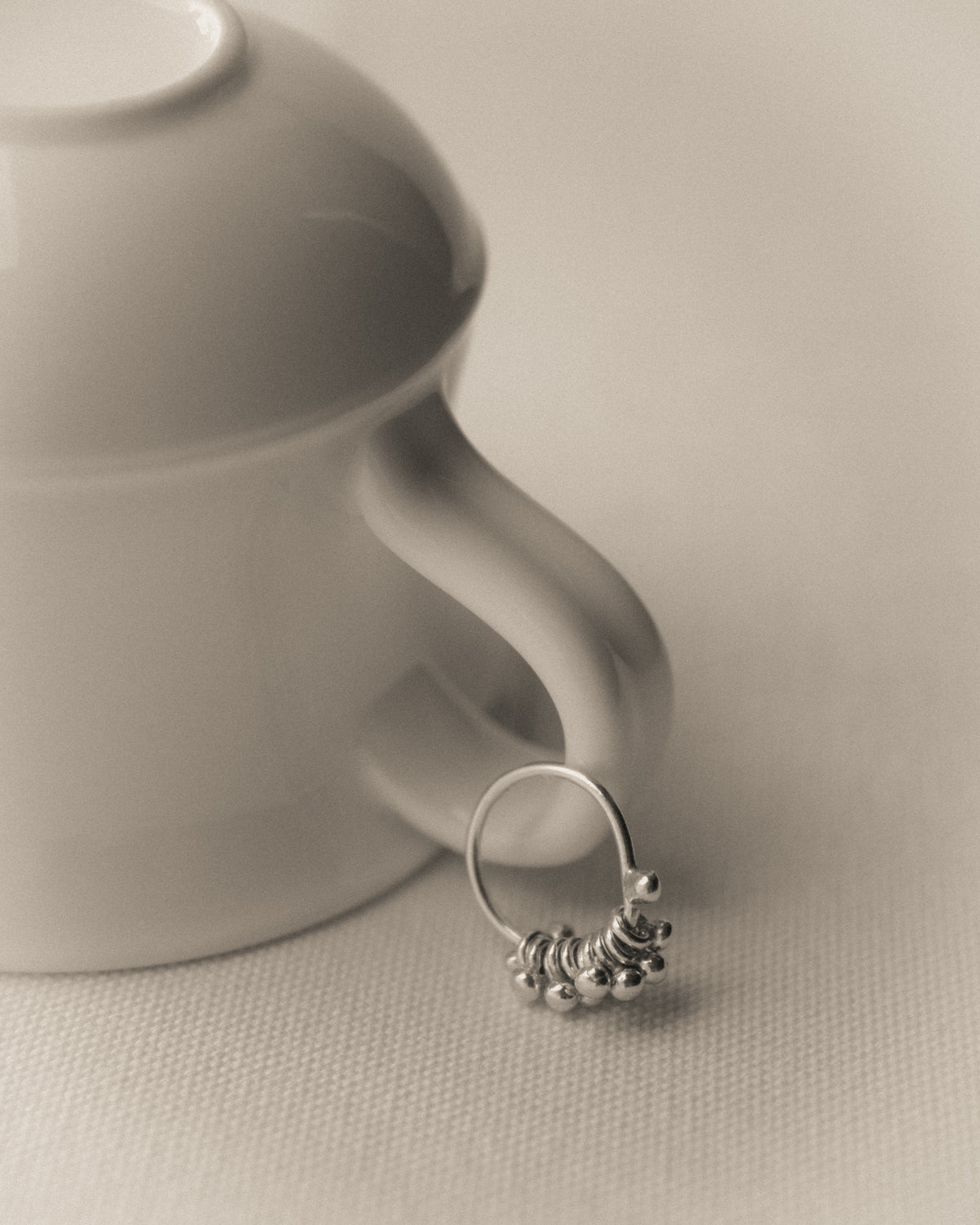 A silver ring featuring a small beaded silver stones design, resting against a white ceramic mug on a textured beige surface.