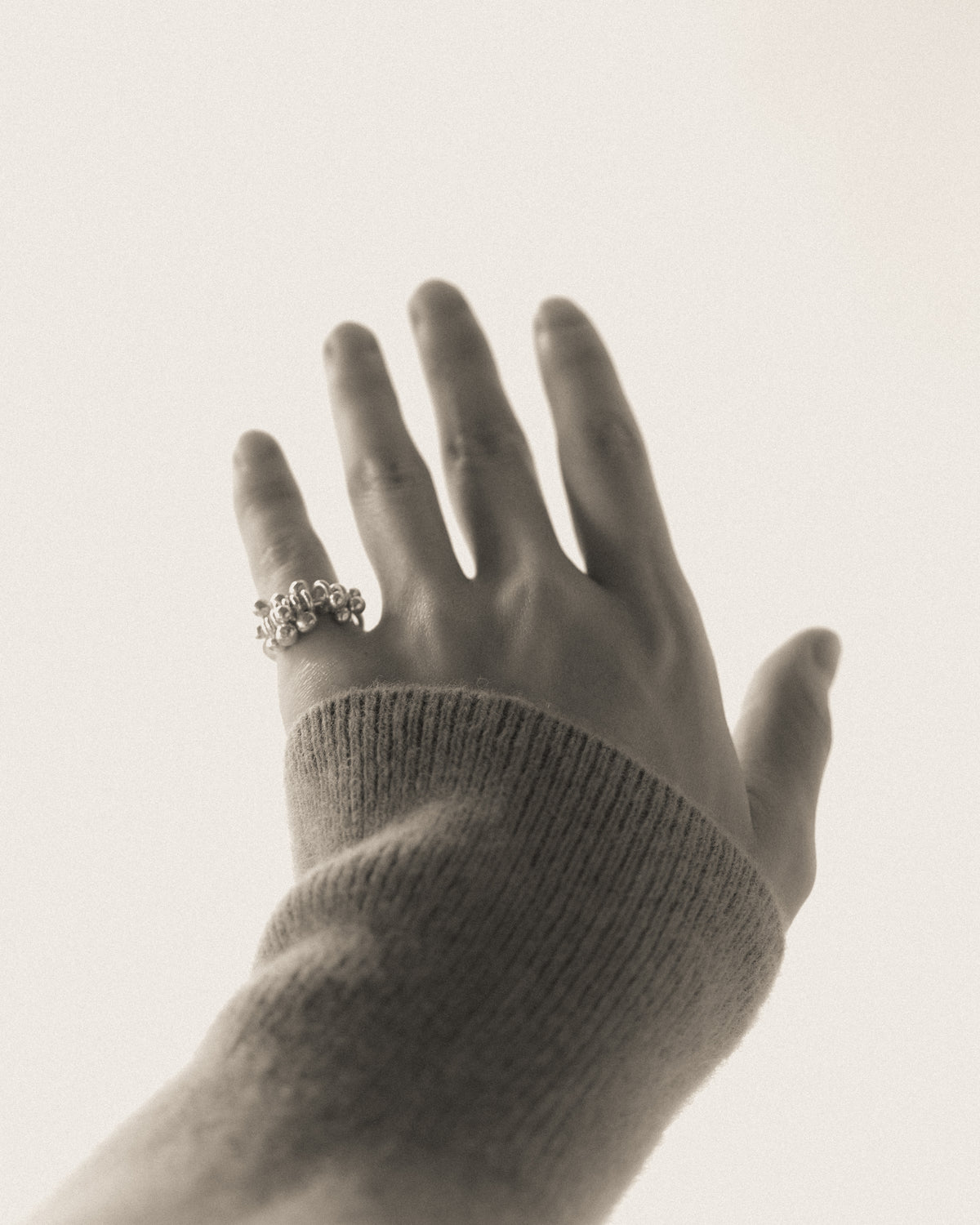 A close-up of a hand wearing a silver ring with small dangling silver stones, shown against a plain light background. The hand is wearing a ribbed knit sweater.