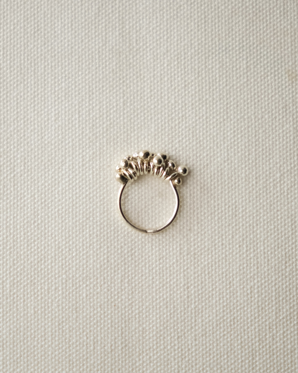 A silver ring with a textured, beaded crown-like top, displayed on a textured off-white fabric background.