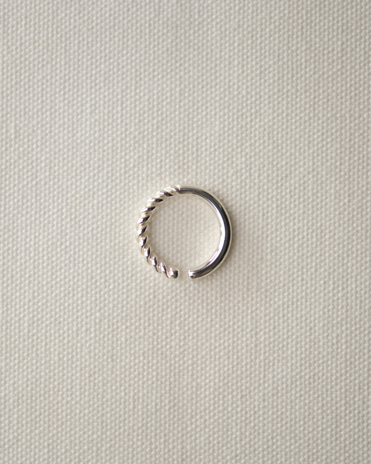 A small silver-toned ring with one twisted segment and one smooth segment, displayed on a textured cream-white fabric background.