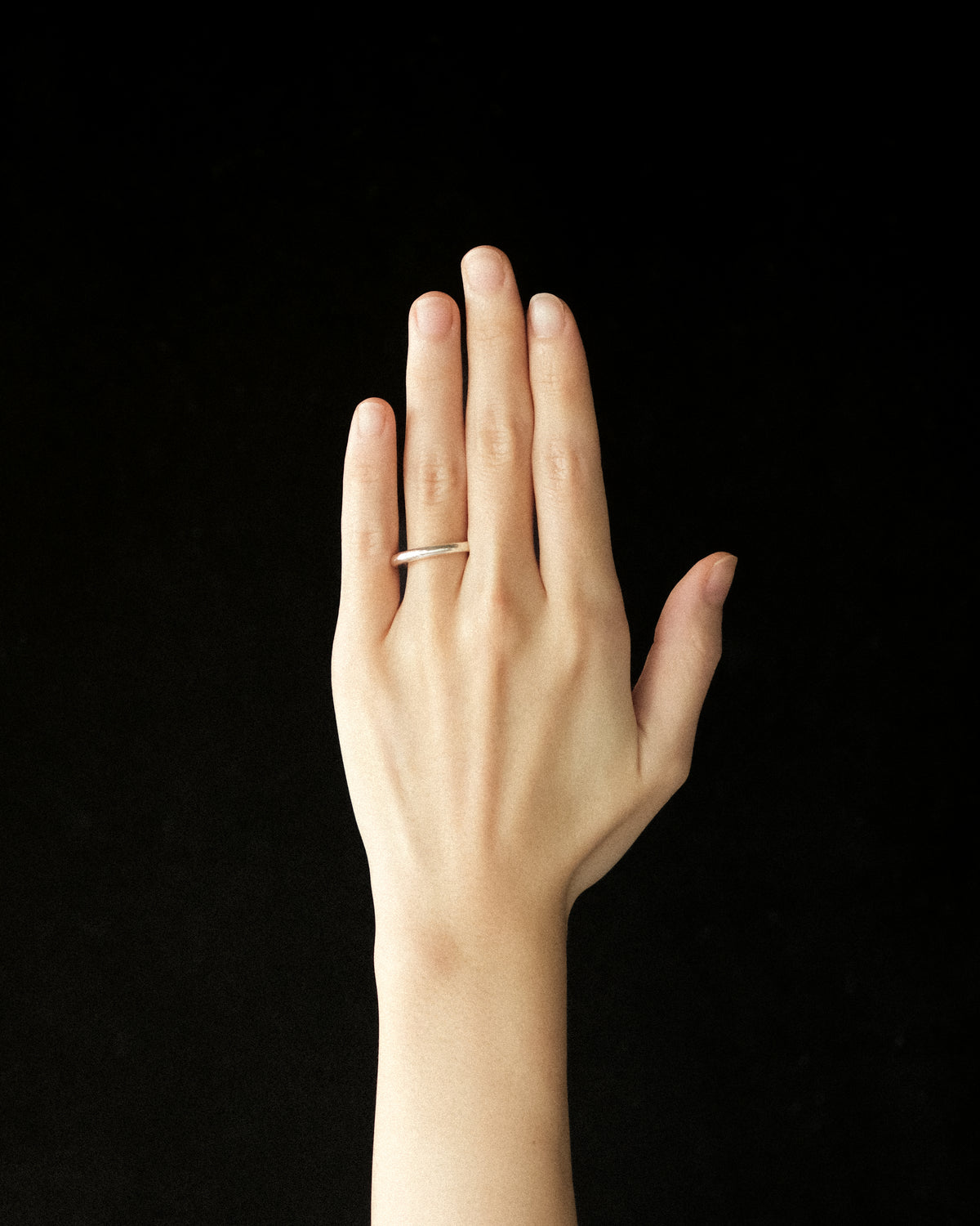 A minimalist silver band ring worn on the ring finger of a hand, shown against a solid black background.