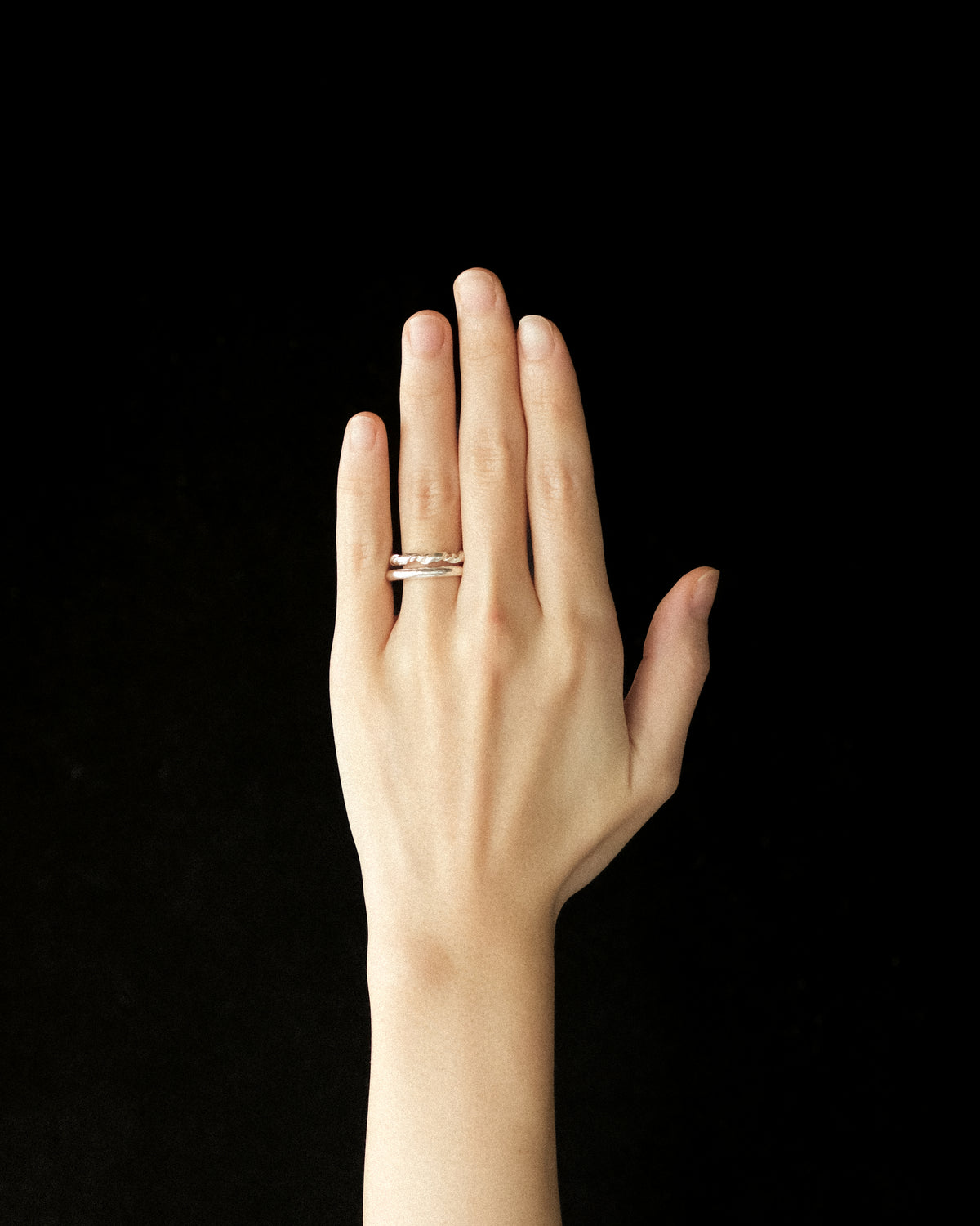 A close-up of a person's hand wearing a minimalist silver band ring stacked with a silver croissant wrap-like ring on the ring finger, set against a solid black background.