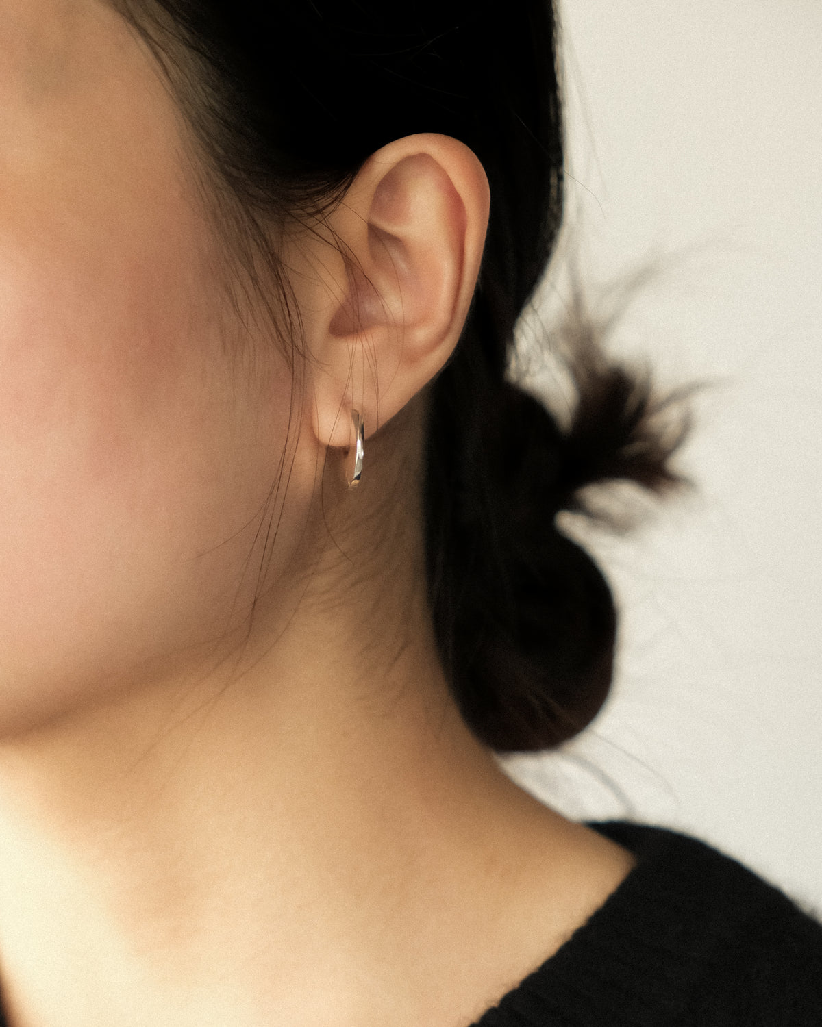 A close-up side profile of a person wearing a small silver huggie hoop earring, with dark hair tied back and a plain light background.