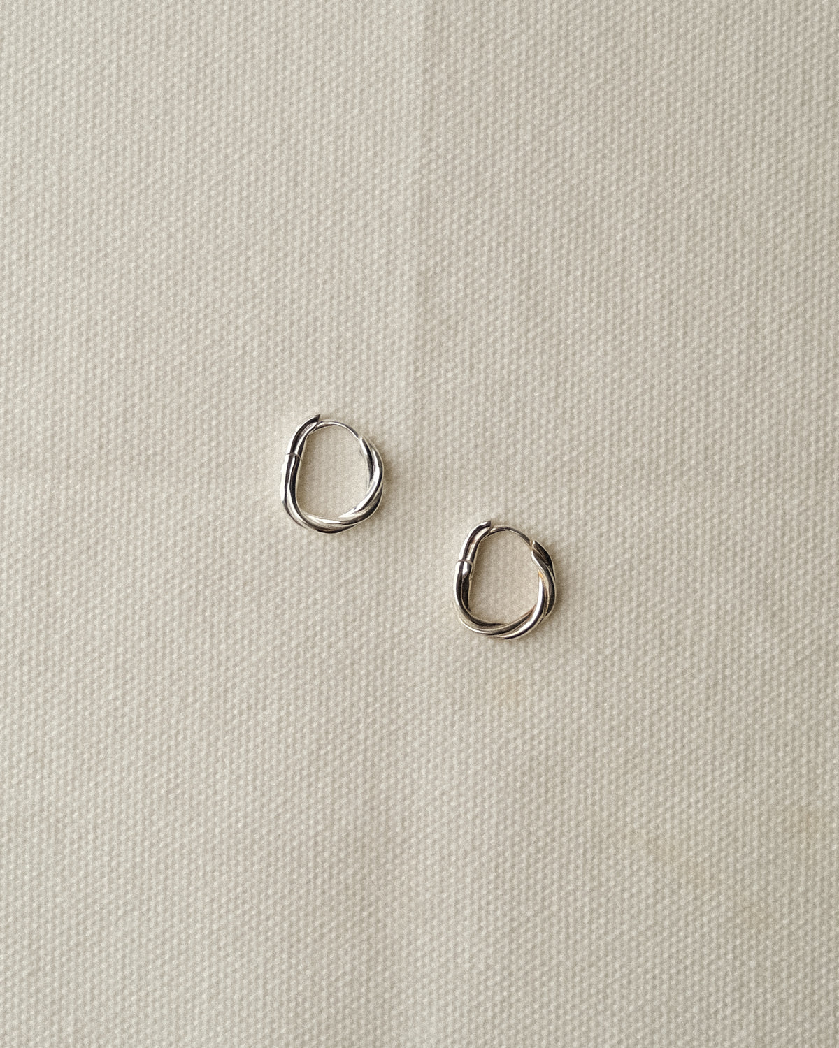 A pair of small silver twisted oval hoop earrings resting on a textured off-white fabric background.