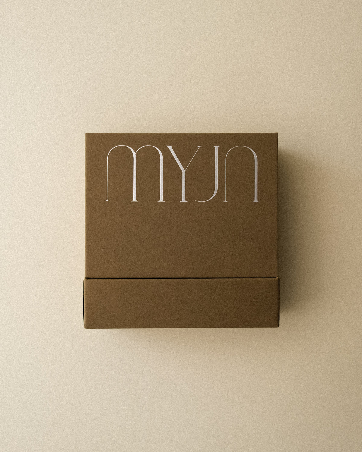 A light khaki brown jewellery box with MYJN branding photographed from above against a warm toned wabi sabi backdrop.