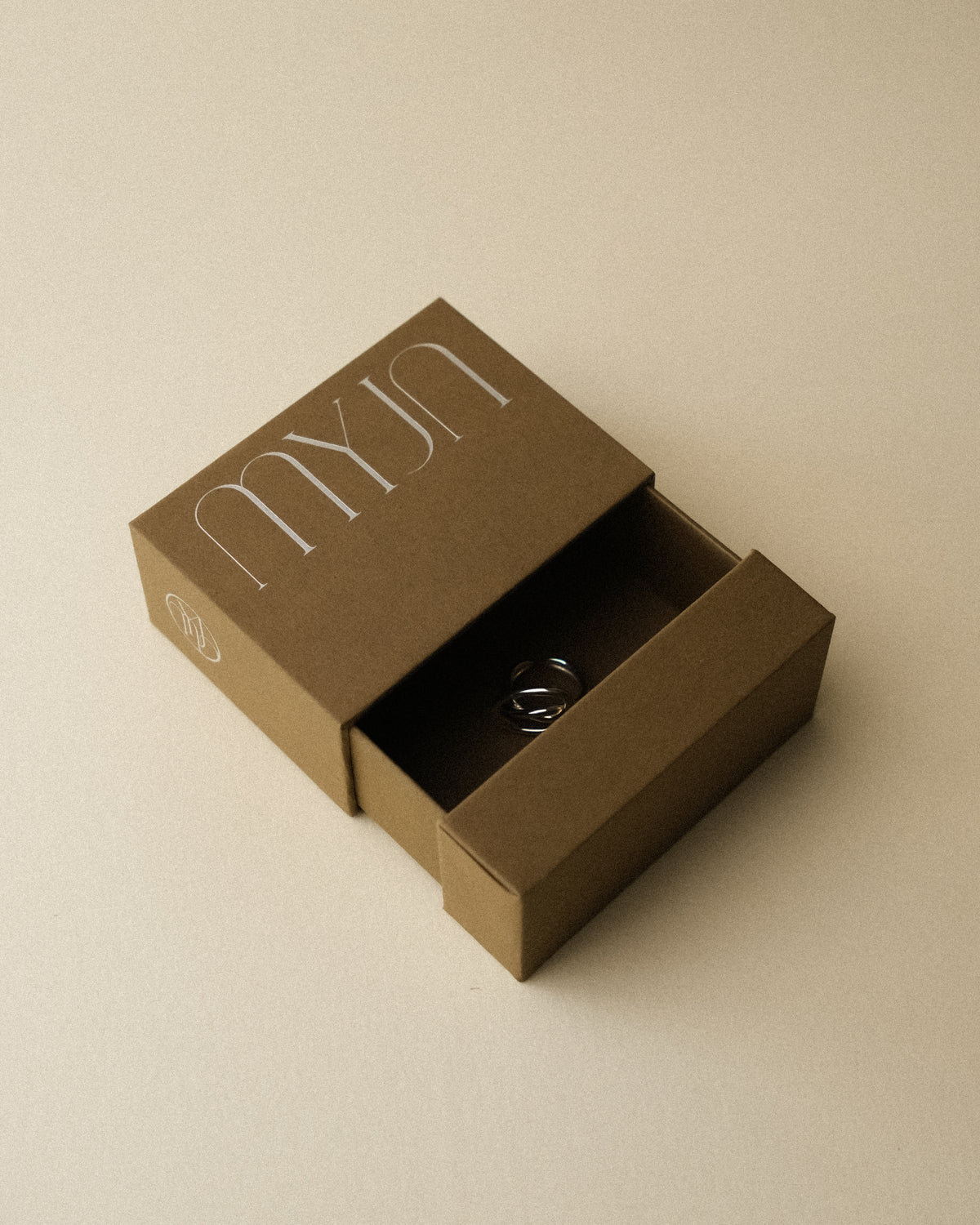 An open light khaki brown jewellery box revealing the silhouette of a ring inside, photographed against a warm toned wabi sabi backdrop.