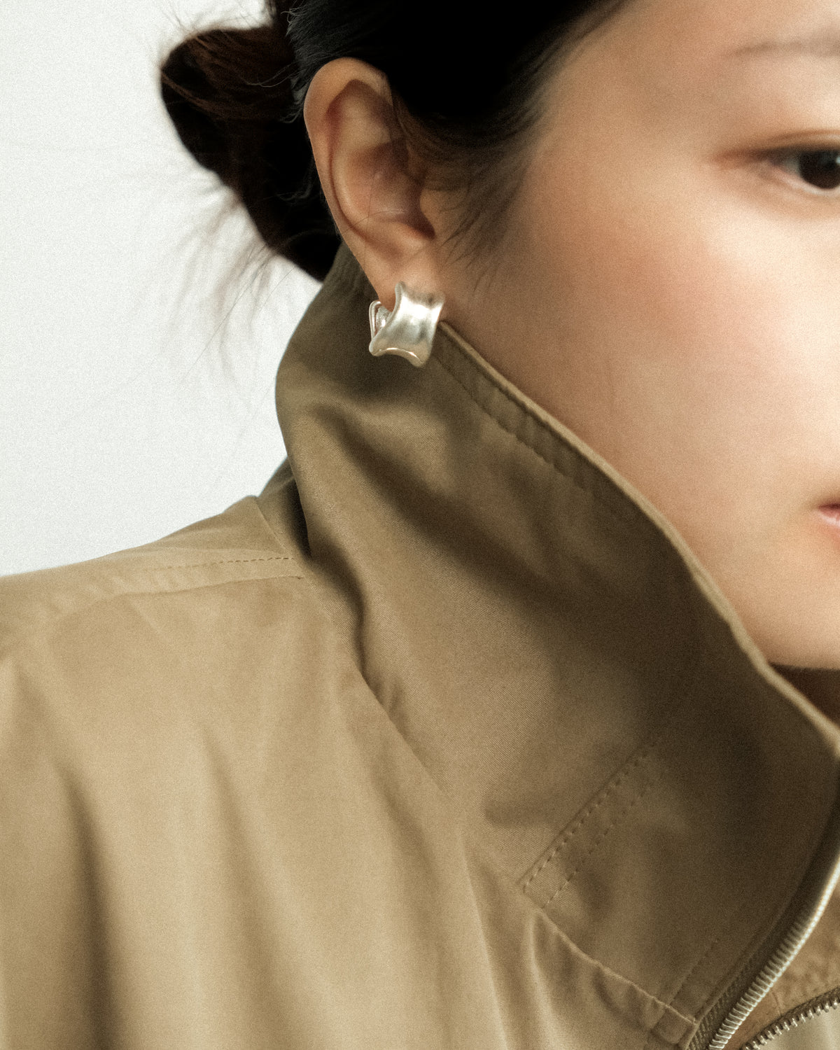 A woman wearing earrings and a high-neck necklace with a khaki coat against white background.