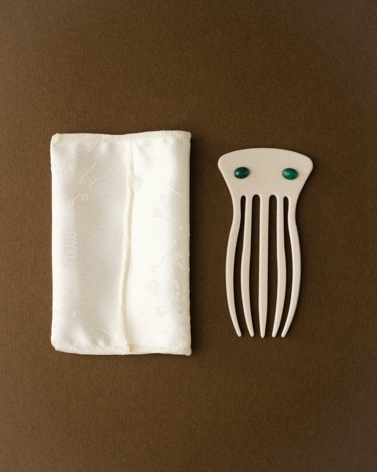 A cream white satin flower print folded pouch paired with an ivory decorative hair comb adorned with two malachite stones, styled against a warm brown backdrop. Soft diffused lighting highlights the satin texture, sculptural form, and rich green malachite accents in a minimal, elegant composition.