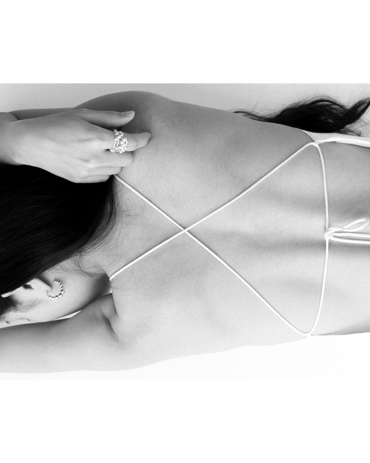 Black and white close-up of a woman lying down from behind, wearing a white string top with thin crisscross back straps and a bold flowing ball ring on one finger with a  silver flowing ball hoop earring.